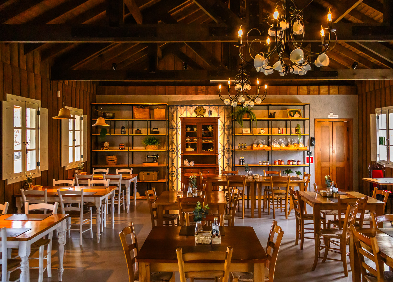 Interior de um restaurante rústico com paredes de madeira, vigas no teto, mesas de jantar preparadas para o serviço e uma área de bar.